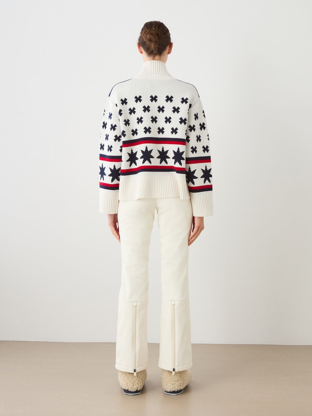 A person stands facing away against a plain backdrop, wearing the We Norwegians EMU x WN Sweater Women in white with navy and red patterns, paired with cream pants featuring back zippers at the hem and beige fuzzy shoes.