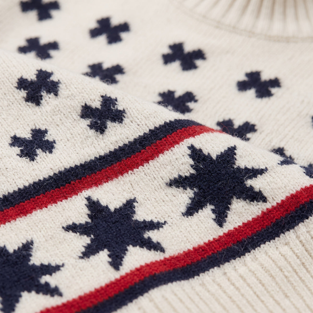 Close-up of the We Norwegians EMU x WN Sweater Women in white, featuring navy blue star and cross patterns with two parallel red and navy stripes near the hem.