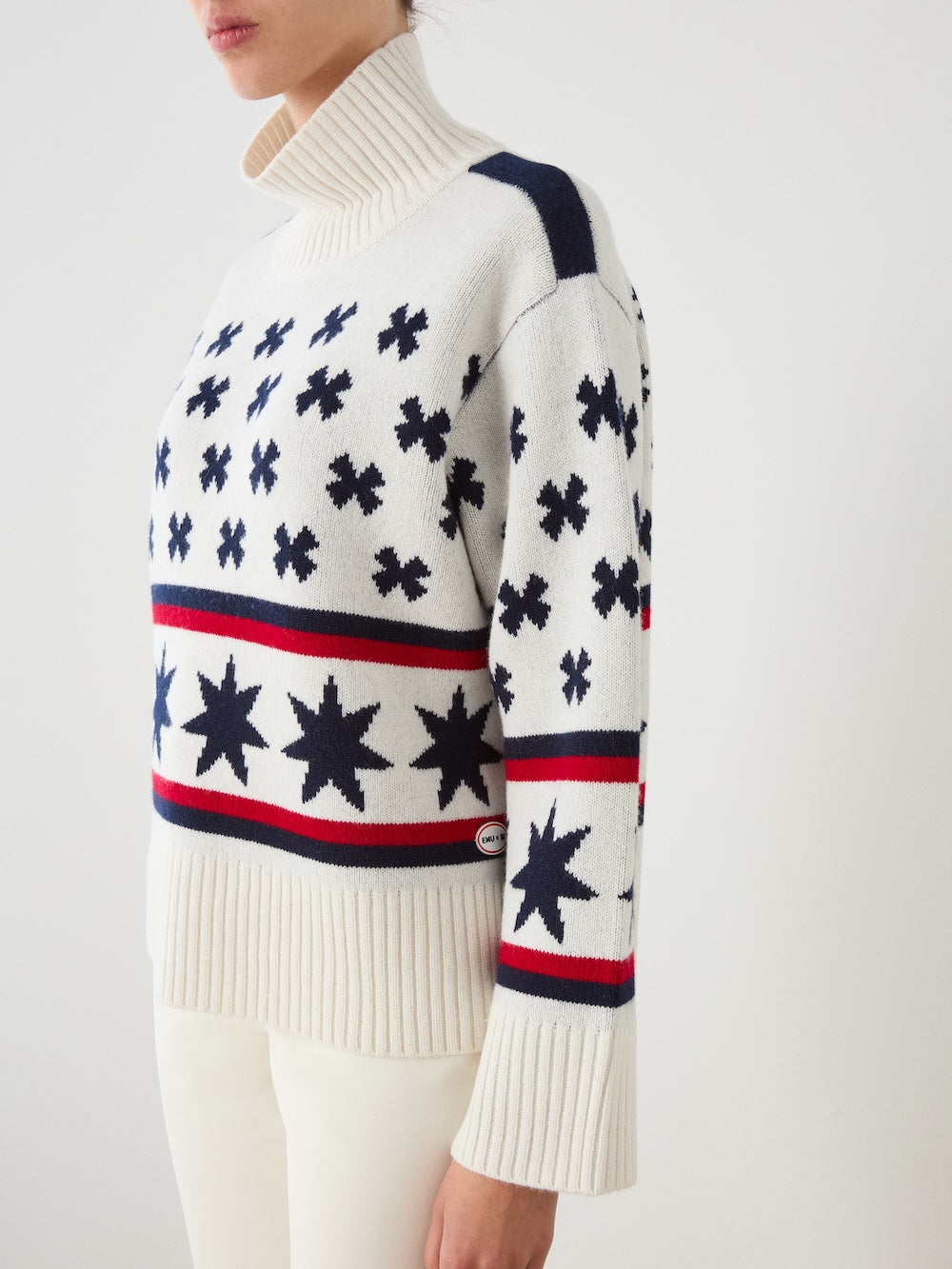 Wearing the We Norwegians EMU x WN Sweater Women—a cozy white turtleneck featuring navy and red star patterns and stripes—paired with white pants, a person stands against a plain light background.