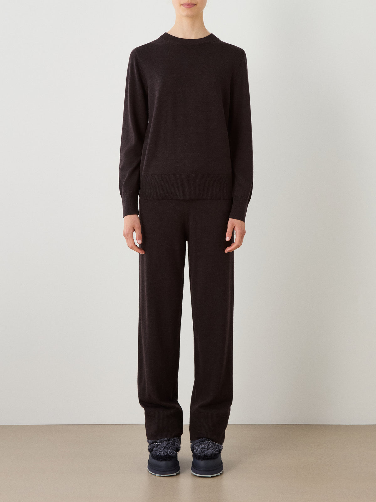 A person stands against a plain light wall, wearing the We Norwegians Geilo Crewneck Women sweater paired with matching loose pants and dark shoes. Their arms rest by their sides, and their face is not visible.