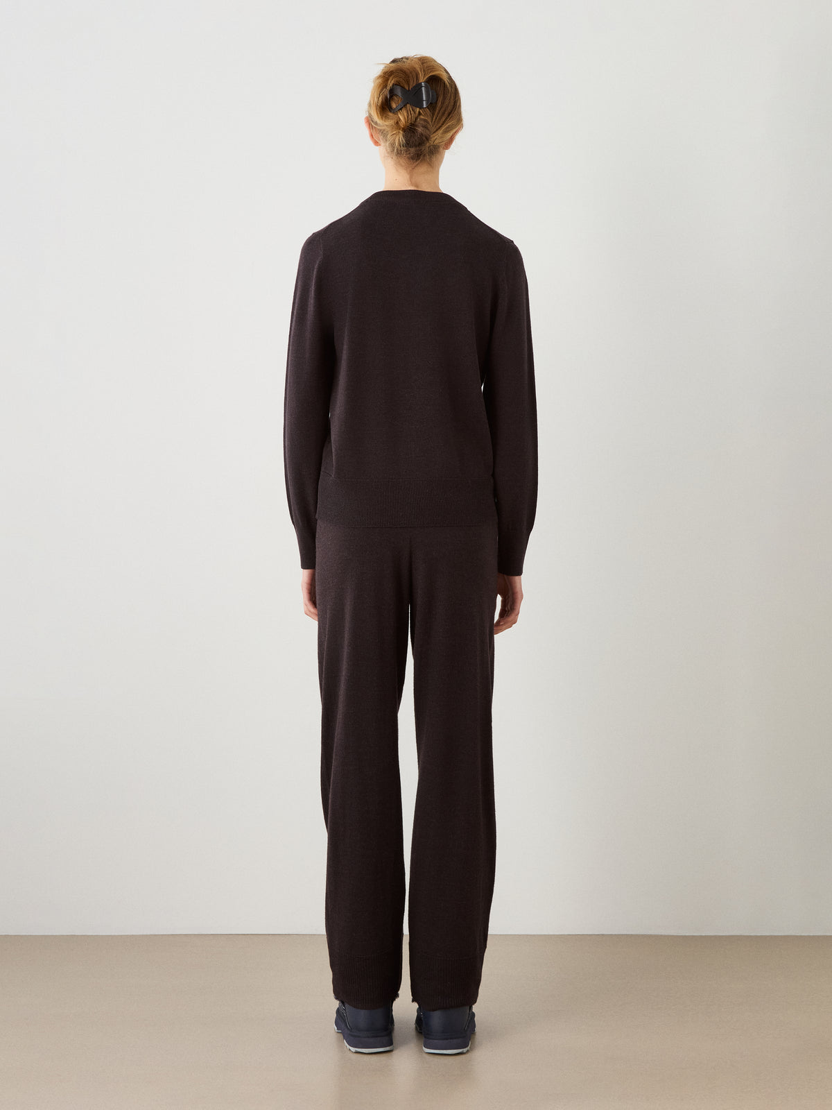 A woman stands indoors facing away from the camera, wearing the We Norwegians Geilo Crewneck sweater paired with matching pants and dark shoes against a plain, minimalistic background.