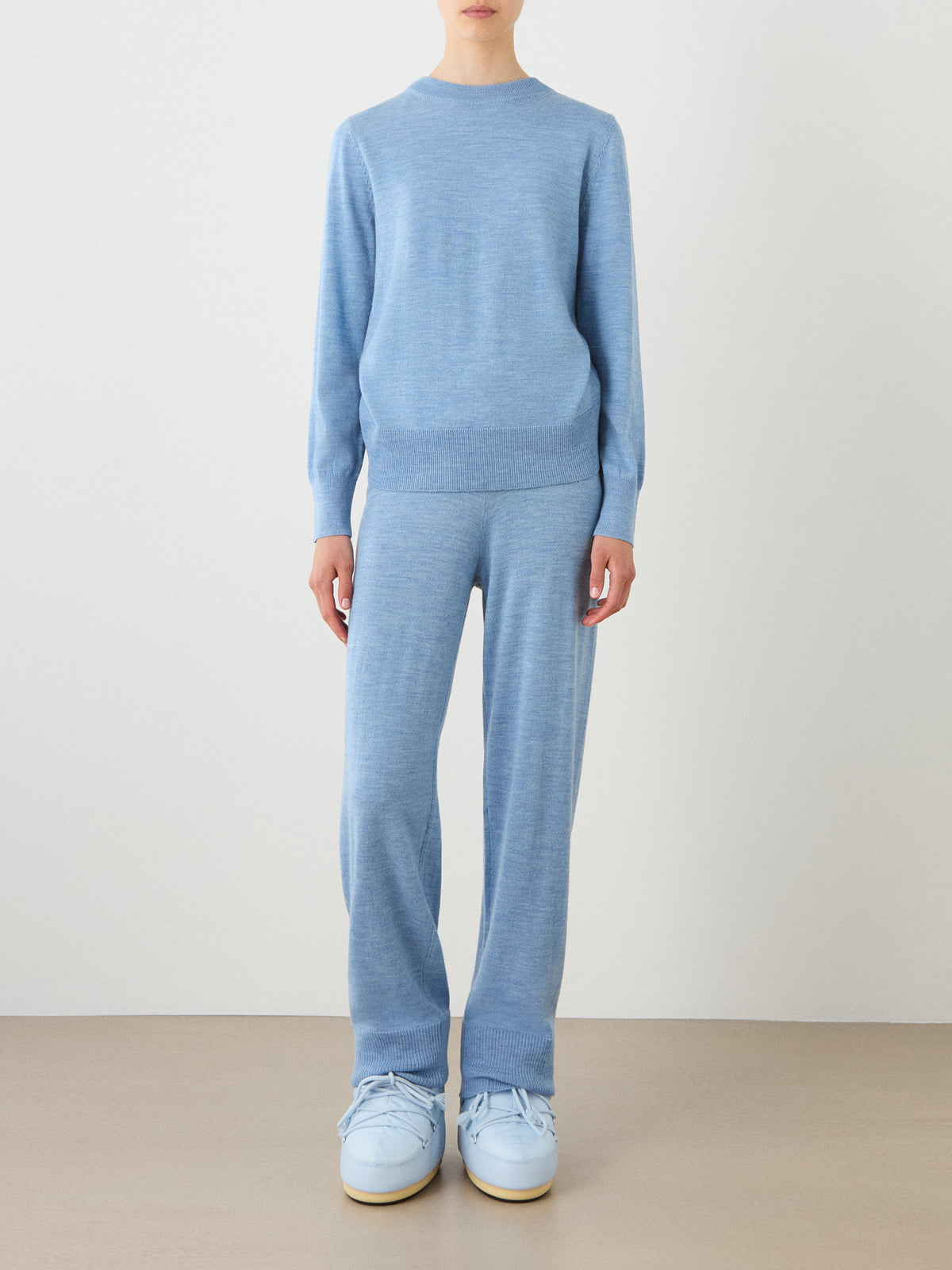 A person is standing against a plain background, wearing the We Norwegians Geilo Crewneck Women in light blue, paired with matching blue pants and oversized light blue sneakers. Their face is not visible in the image.