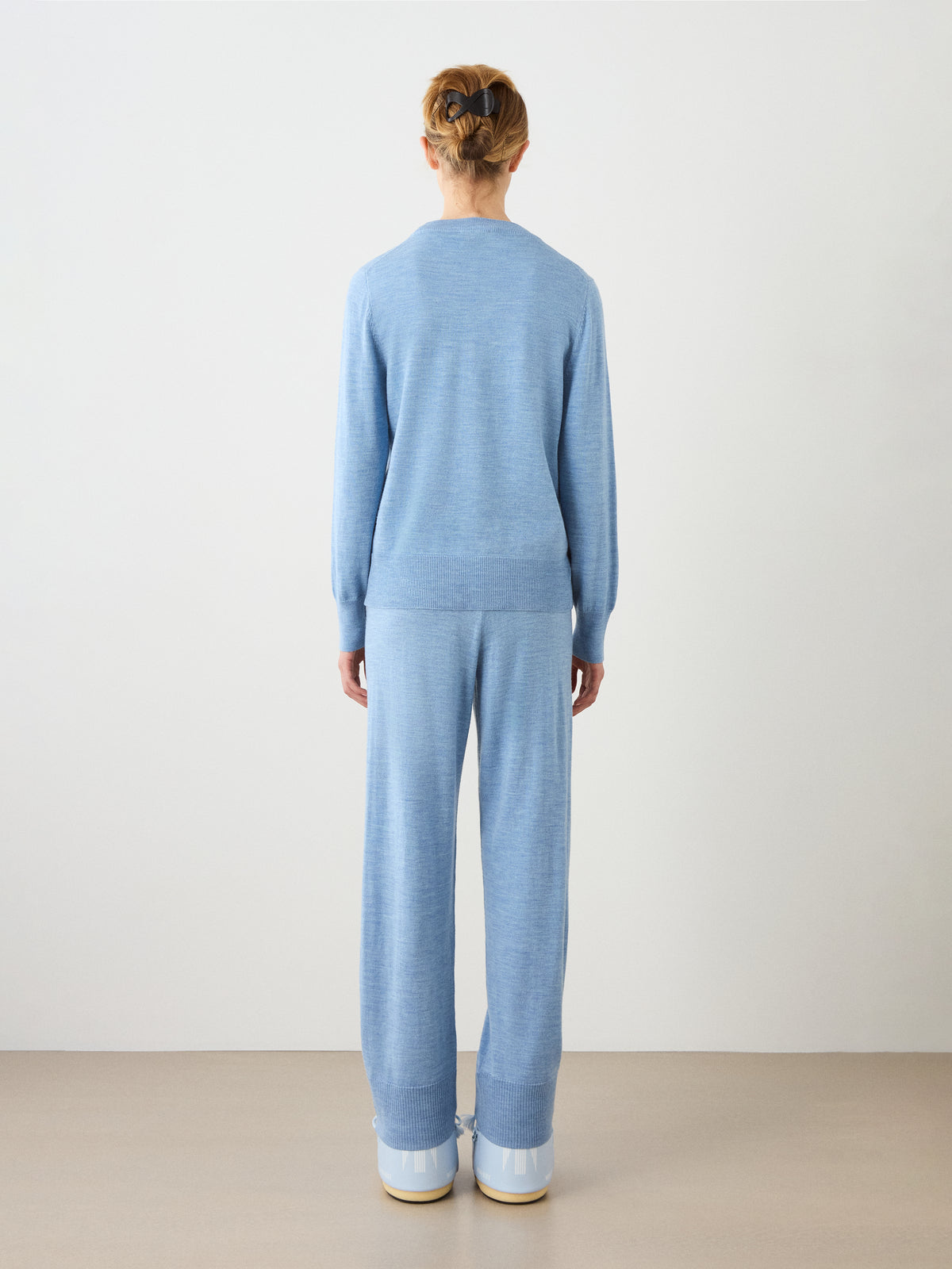 A person stands facing away from the camera, wearing the We Norwegians Geilo Crewneck Women sweater in light blue with matching wide-leg pants and white shoes, in a minimal studio setting with a plain background.