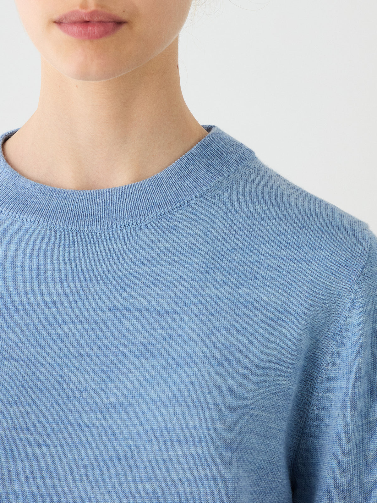 A close-up of a person wearing the We Norwegians Geilo Crewneck Women sweater in light blue, with the lower half of their face and neck visible against a plain background.