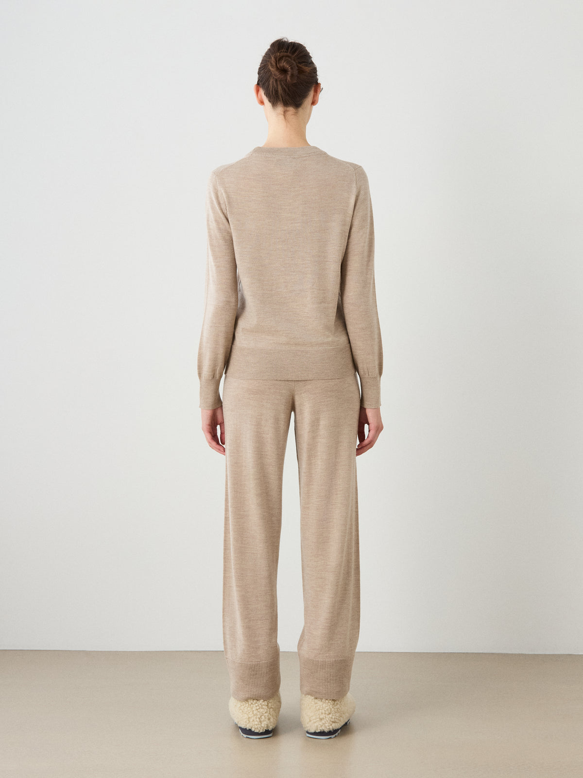 A person stands facing away, wearing the We Norwegians Geilo Crewneck Women sweater in beige as part of a luxury lounge set with matching wide-leg pants and cream-colored slippers. The background is plain and minimalistic.