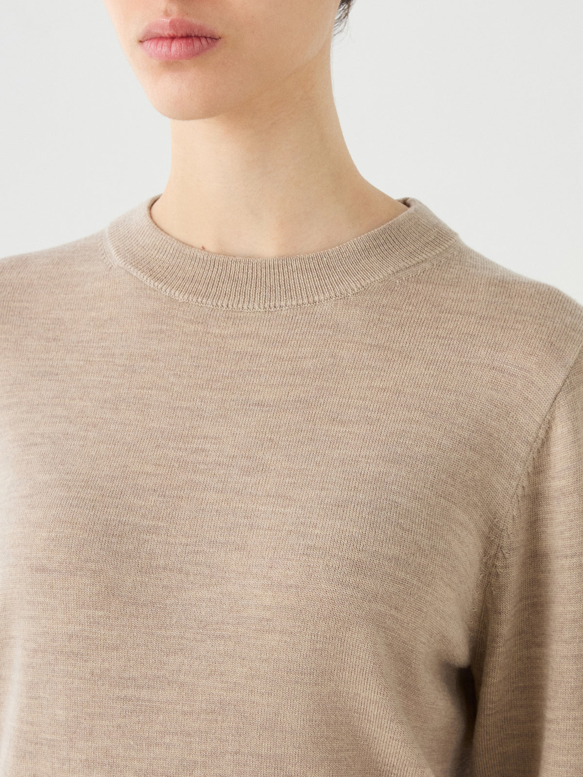 A person is shown from the shoulders to just above the lips against a plain light background, wearing the We Norwegians Geilo Crewneck Women—a light beige, long-sleeve merino wool sweater.