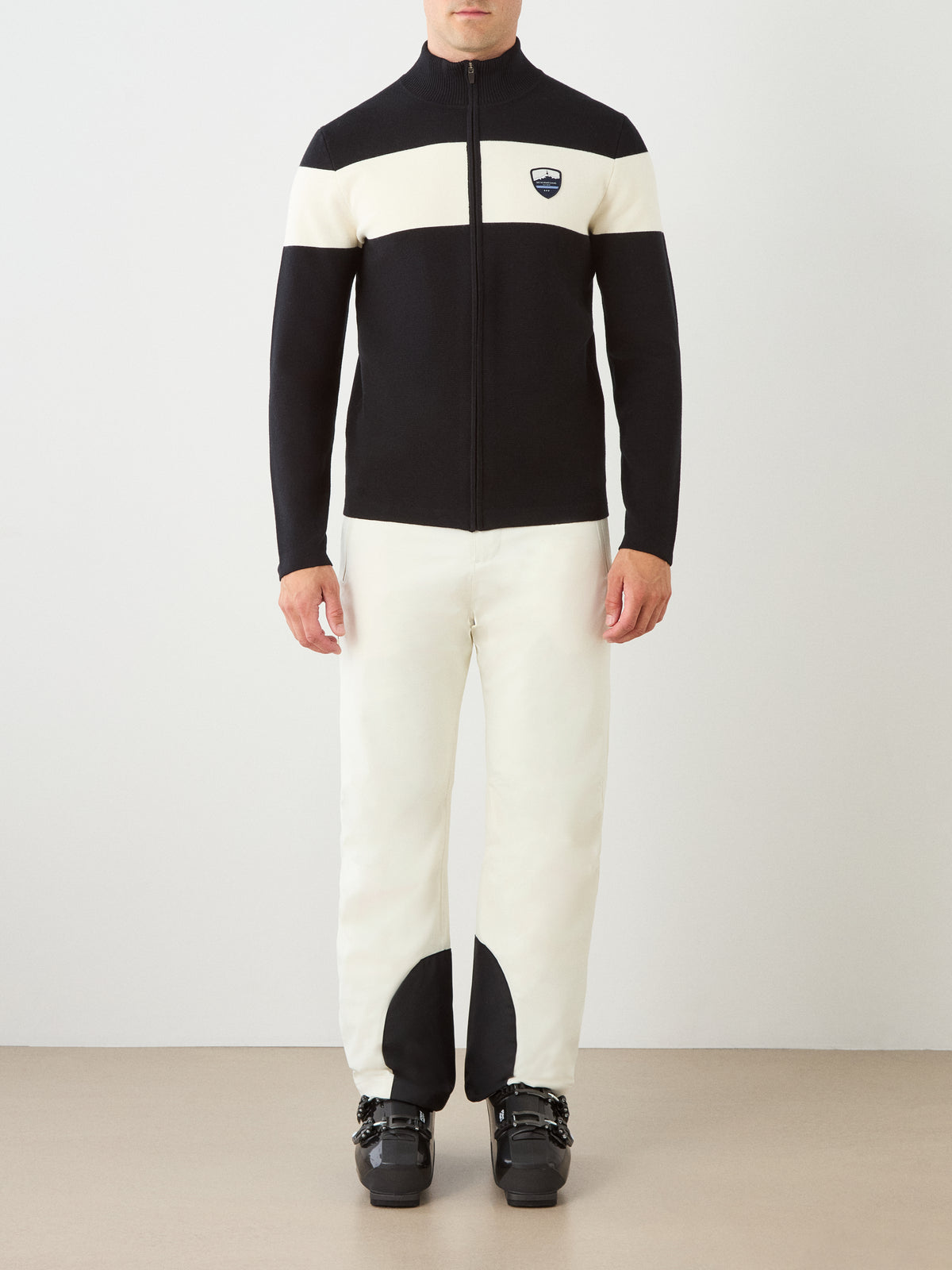 A man stands against a plain background wearing the We Norwegians Holmenkollen Full Zip Men jacket in black and white, along with white pants featuring black patches at the ankles and black ski boots—a look inspired by Scandinavian mountain heritage. His face is not visible.
