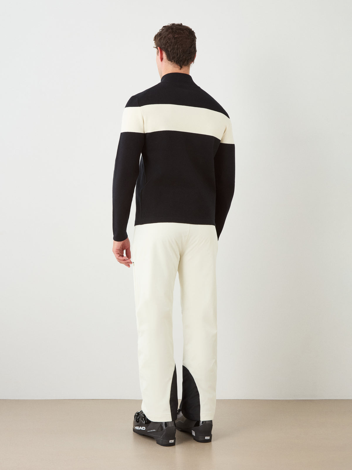 A person stands facing away, dressed in the We Norwegians Holmenkollen Full Zip Men merino wool sweater in black with a white horizontal stripe, paired with off-white pants and black boots against a plain background—a look inspired by Scandinavian mountain heritage.