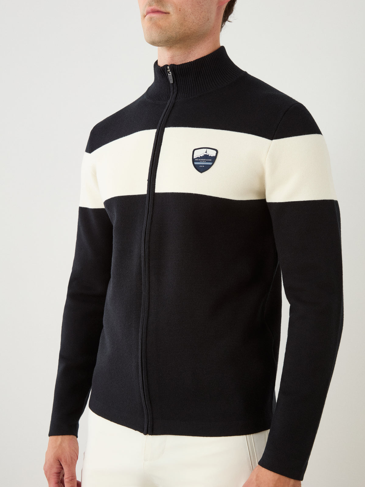 A man wears the Holmenkollen Full Zip Men merino wool sweater by We Norwegians in black, featuring a white horizontal stripe across the chest and a small shield logo on the left side, paired with white pants—an ensemble that captures classic Scandinavian mountain heritage.