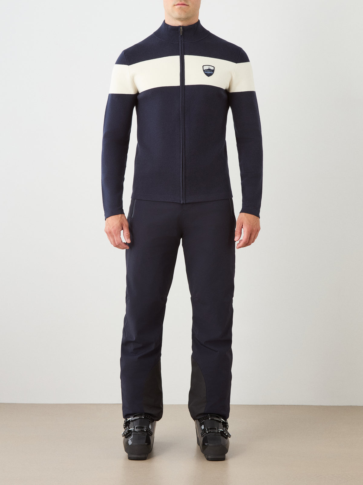 A man stands facing forward against a neutral background, wearing the We Norwegians Holmenkollen Full Zip Men jacket—a navy layering piece with a white horizontal stripe—over a merino wool sweater, paired with dark pants and black ski boots.