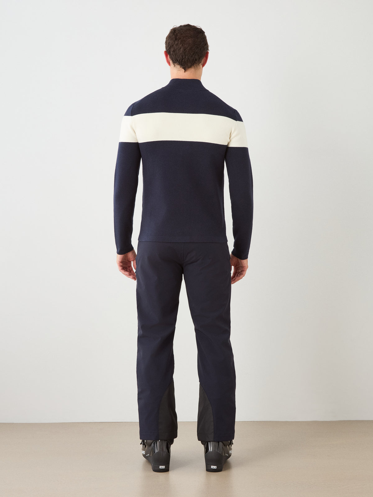 A person stands facing away, wearing the We Norwegians Holmenkollen Full Zip Men sweater—a navy merino wool piece featuring a white horizontal stripe across the back, inspired by Scandinavian mountain heritage—paired with dark pants and black boots, against a plain light background.