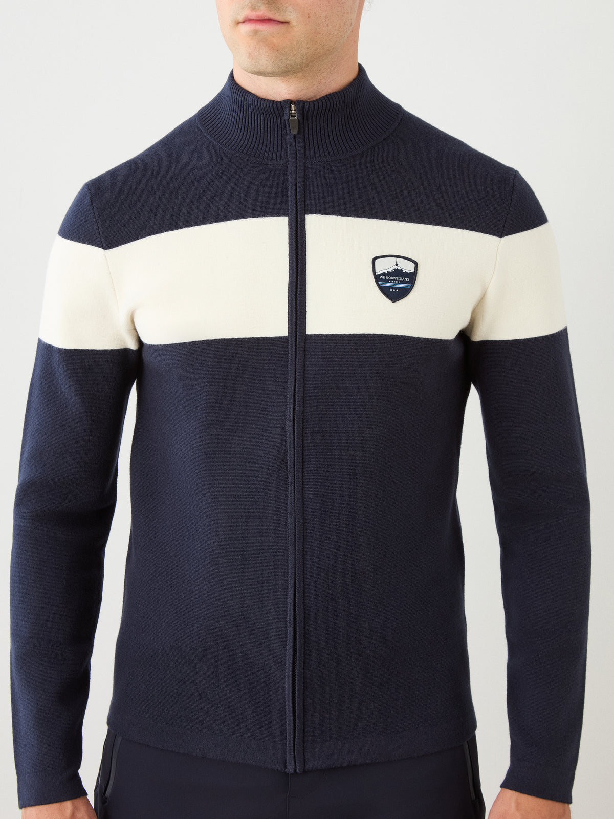 A man wearing the We Norwegians Holmenkollen Full Zip Men jacket in navy, distinguished by a white horizontal stripe across the chest and arms and adorned with a subtle shield-shaped logo on the left chest—an understated tribute to Scandinavian mountain heritage.