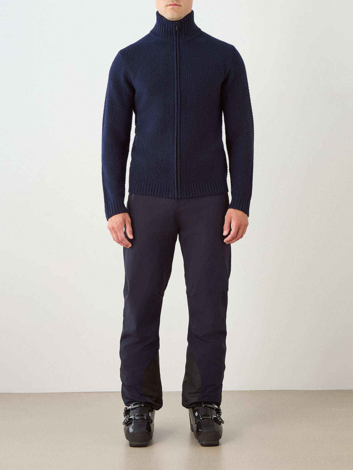 A person stands against a plain background wearing the We Norwegians Kikut FullZip Men sweater, styled as a full-zip cardigan in navy, paired with black pants and black ski boots.