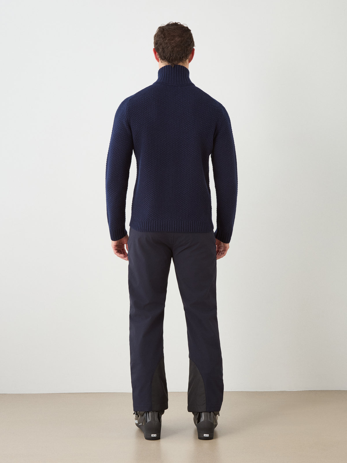 A man stands facing away, wearing the We Norwegians Kikut FullZip Men navy alpine sweater paired with black pants and black ski boots, set against a plain light background.
