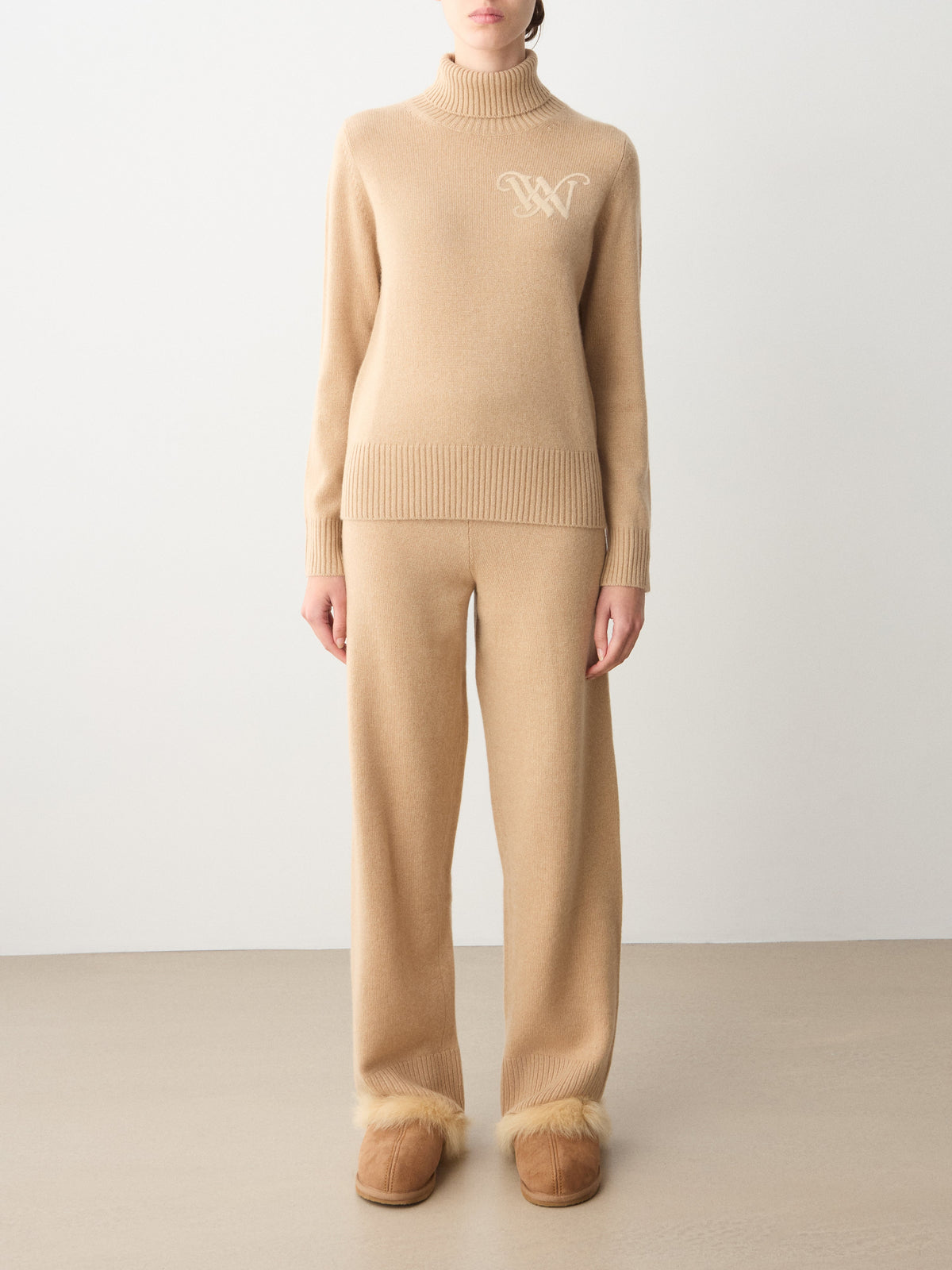 A person wearing a beige We Norwegians Monogram Cashmere Sweater Women with a chest monogram, matching wide-leg trousers, and fur-trimmed slippers in an ensemble of luxury loungewear.