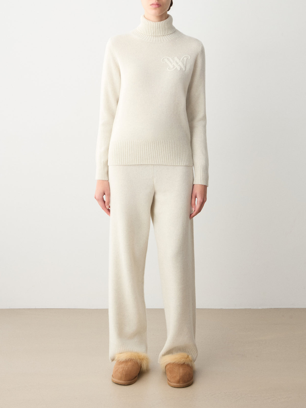 A person wearing a cream-colored We Norwegians Monogram Cashmere Sweater Women with an embroidered monogram on the chest, matching lounge pants, and tan fur-lined slippers.