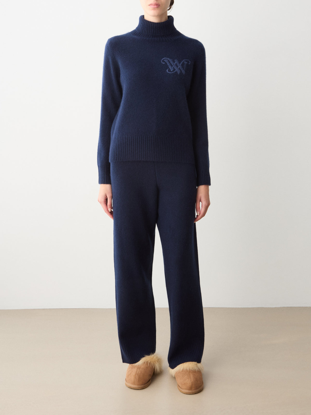 A person wearing a navy blue Monogram Cashmere Sweater Women with a tonal monogram on the chest, matching pants, and tan shearling-lined slippers.