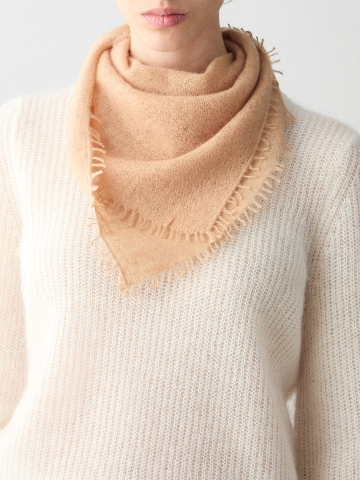 A light tan Monogram Cashmere Scarf by We Norwegians, styled as a featherlight triangular scarf with frayed edges, worn around the neck over a white knit sweater.