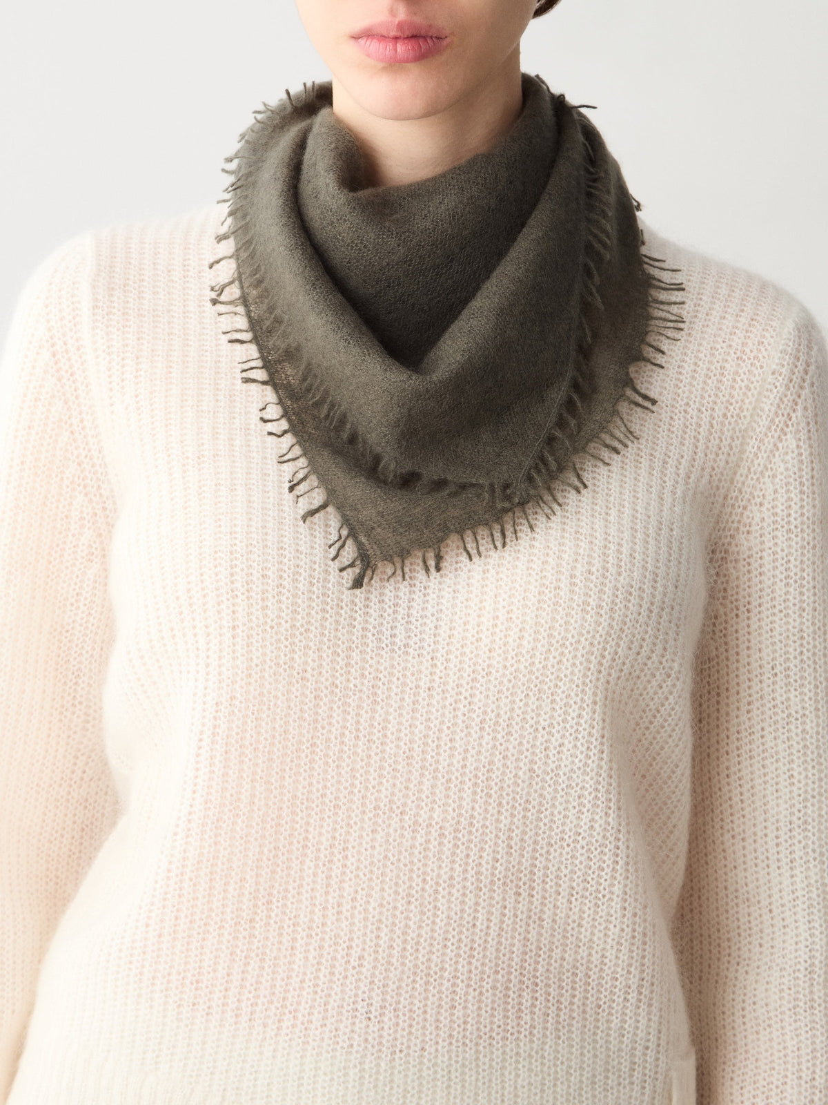 A person wearing a dark olive Monogram Cashmere Scarf in a featherlight, triangle shape with a fringed edge over a white knit sweater.