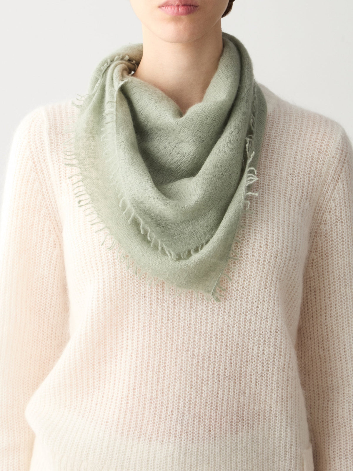 Sage green, featherlight Monogram Cashmere Scarf in a triangular shape with frayed edges draped around a person’s neck.