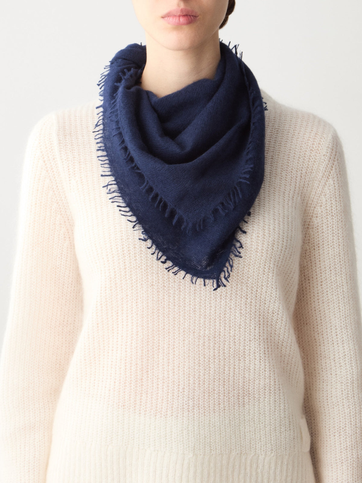 A person wearing a featherlight navy blue triangular Monogram Cashmere Scarf with frayed edges over a cream-colored sweater.