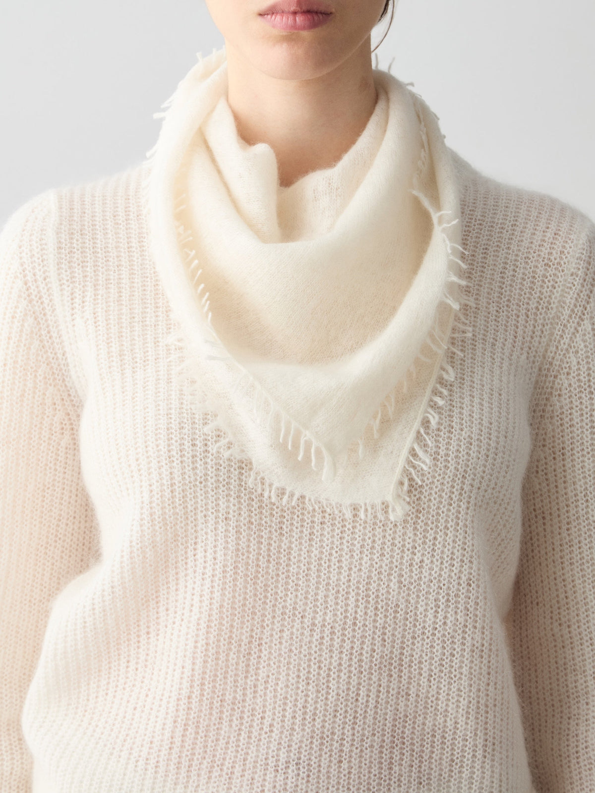 A person wearing a featherlight, cream-colored triangular Monogram Cashmere Scarf with frayed edges over a matching cashmere sweater.