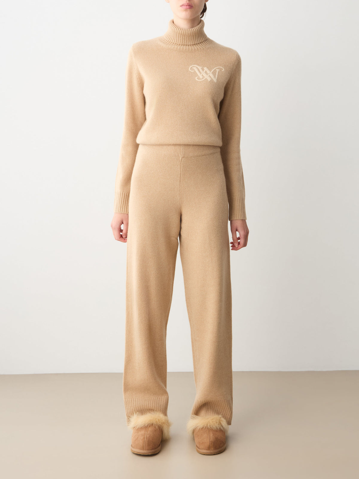 A person wearing a beige We Norwegians Monogram Jogger Women turtleneck sweater and matching wide-leg cashmere pants, styled with tan fur-lined slippers for comfortable travel wear.