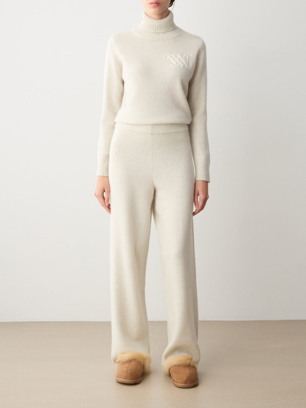 A person wearing a cream-colored turtleneck sweater with a white monogram and matching We Norwegians Monogram Jogger Women trousers, paired with brown fur-lined slip-on shoes.