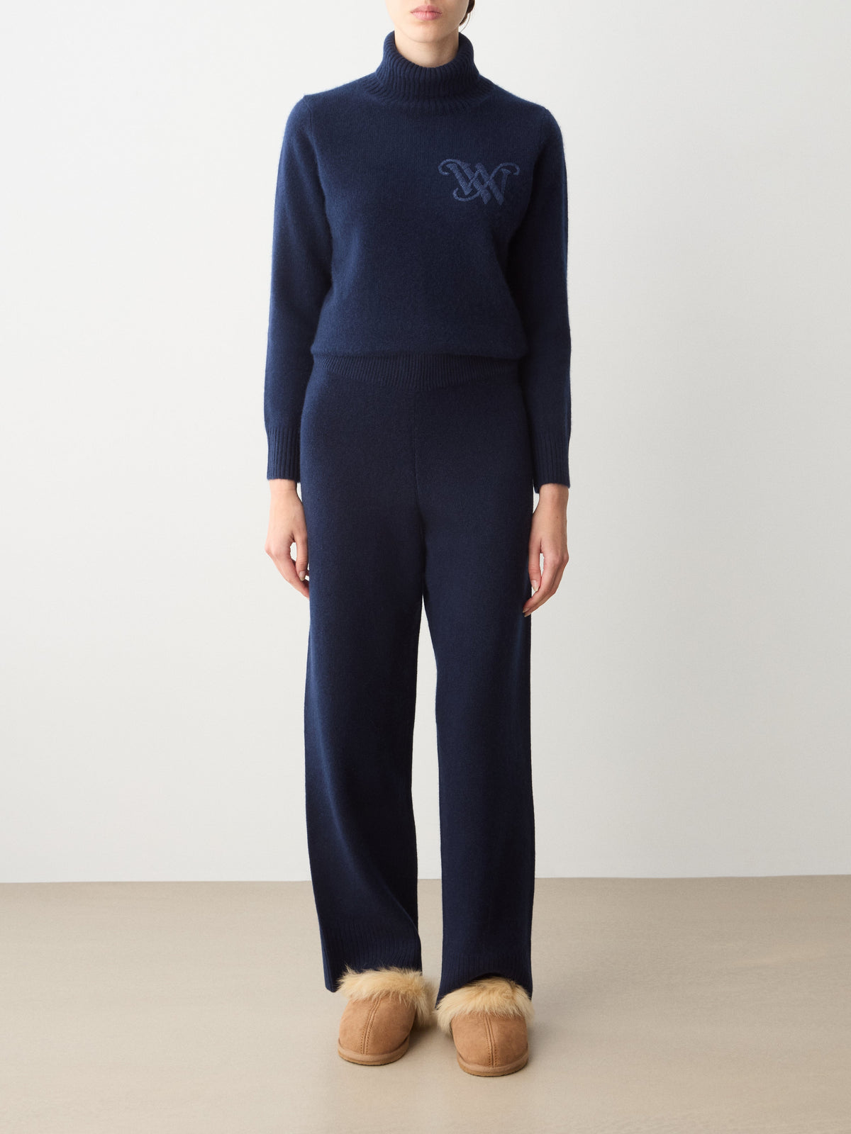 A person wearing a navy blue turtleneck sweater and matching We Norwegians Monogram Jogger Women cashmere pants, paired with tan fur-lined slip-on shoes for comfortable travel wear.