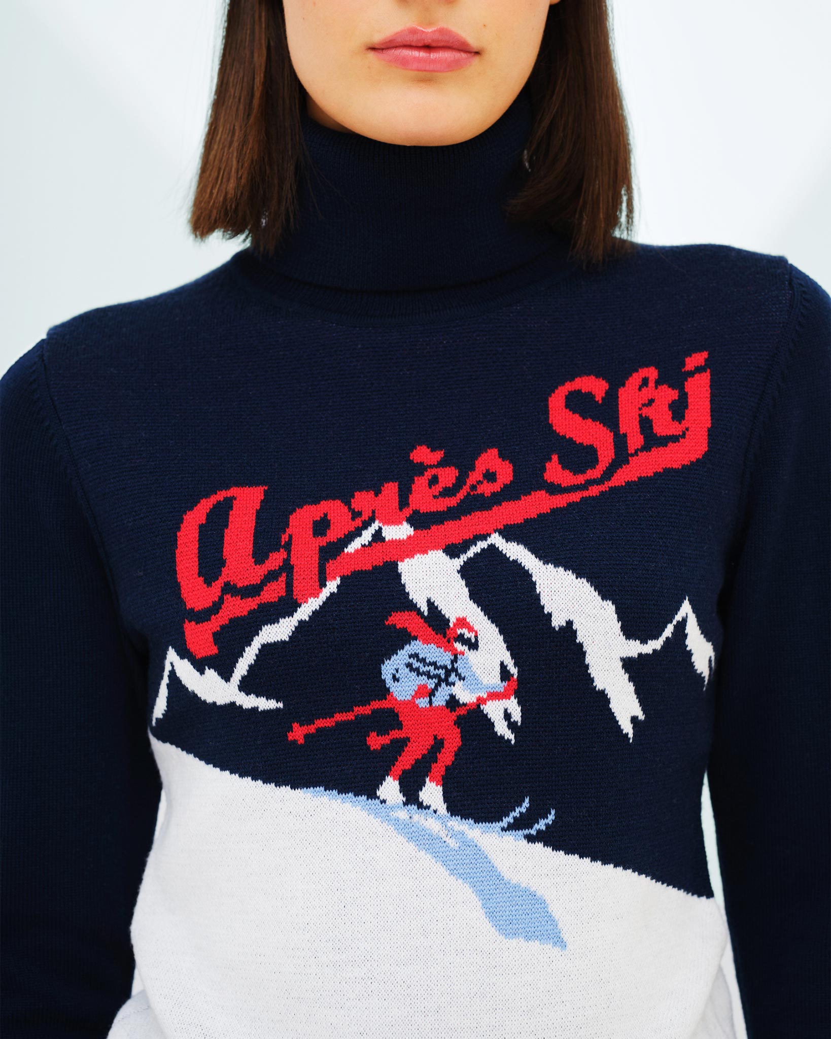 A woman is wearing the Afterski Sweater Women by We Norwegians, a navy blue turtleneck made of Merino wool. It features an embroidered design of a skier and snowy mountains, perfectly capturing the après ski look, with the text "Après Ski" written above the skier in red.