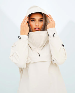 A woman wearing a light beige Beito Anorak Women by We Norwegians looks directly at the camera, holding the sides of the hood with both hands. The background is a light, minimalist setting.