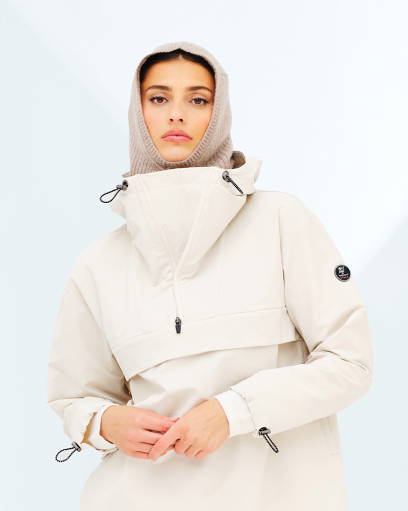 A person wearing a light beige anorak with a high collar and a hood, over what appears to be the sophisticated Blefjell Balaclava Women by We Norwegians, made from knitted beige fabric in a 1/2 cardigan stitch. The jacket features pull cords on the collar and cuffs and bears a black round logo on the left sleeve. The background is light with subtle geometric shadows.