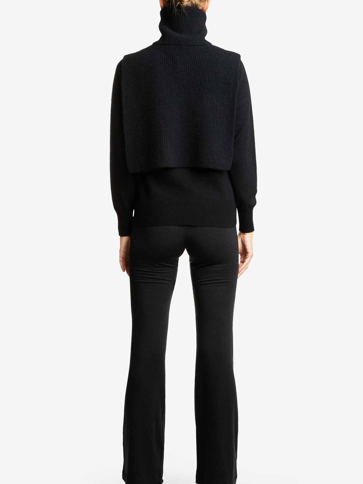 A person is seen from behind, highlighting the Blefjell Bib Women by We Norwegians, a chic black turtleneck sweater with a designer vest layered panel. It is tastefully paired with black flared pants and complemented by a light bun hairstyle. The minimalist white background enhances the modern elegance of the outfit.