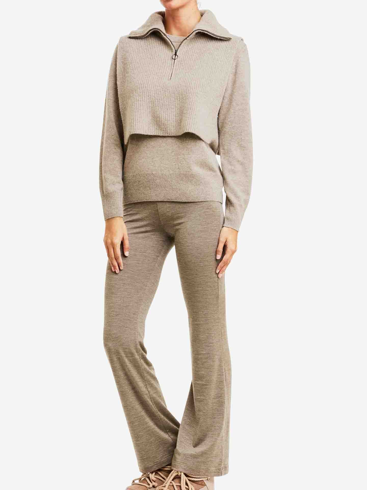 A person is dressed in the Blefjell Cashmere Bib Women by We Norwegians, featuring a light brown knitted sweater with a short cashmere bib overlay, complemented by matching flared pants and beige shoes. The background is plain white.