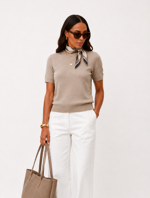 A woman wearing sunglasses, the We Norwegians Club Top Women – Summer Cashmere in beige, white wide-leg pants, taupe flats, and a patterned scarf carries a large taupe tote bag while standing against a plain white background.