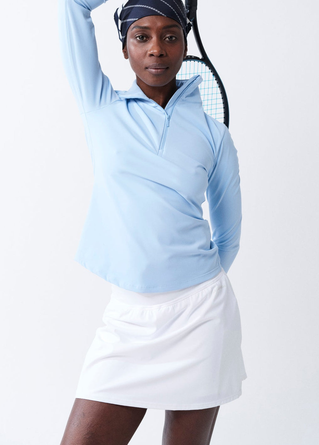 Person wearing a light blue long-sleeve top and white skirt holding a tennis racket on a white background