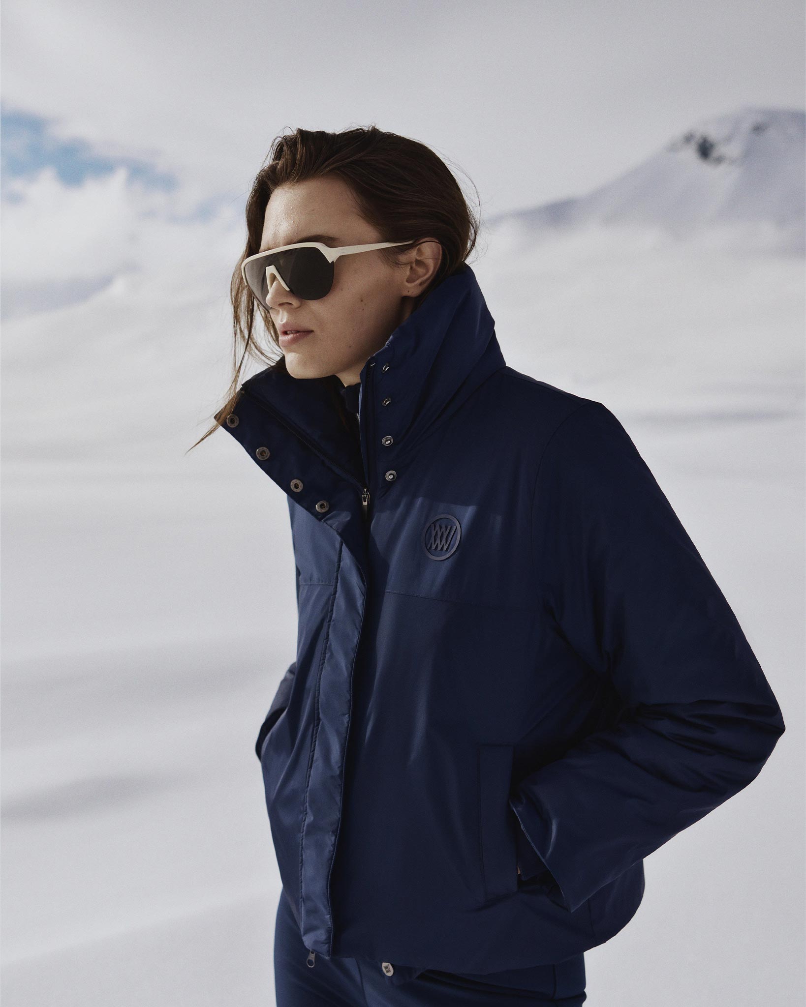 A person wearing the We Norwegians Filefjell Ski Jacket Women, which features wool padding and is navy blue, strides through a snowy landscape with large sunglasses on. The background showcases snow-covered hills beneath a cloudy sky. The jacket has a high collar, and the person casually rests their hands in their pockets.