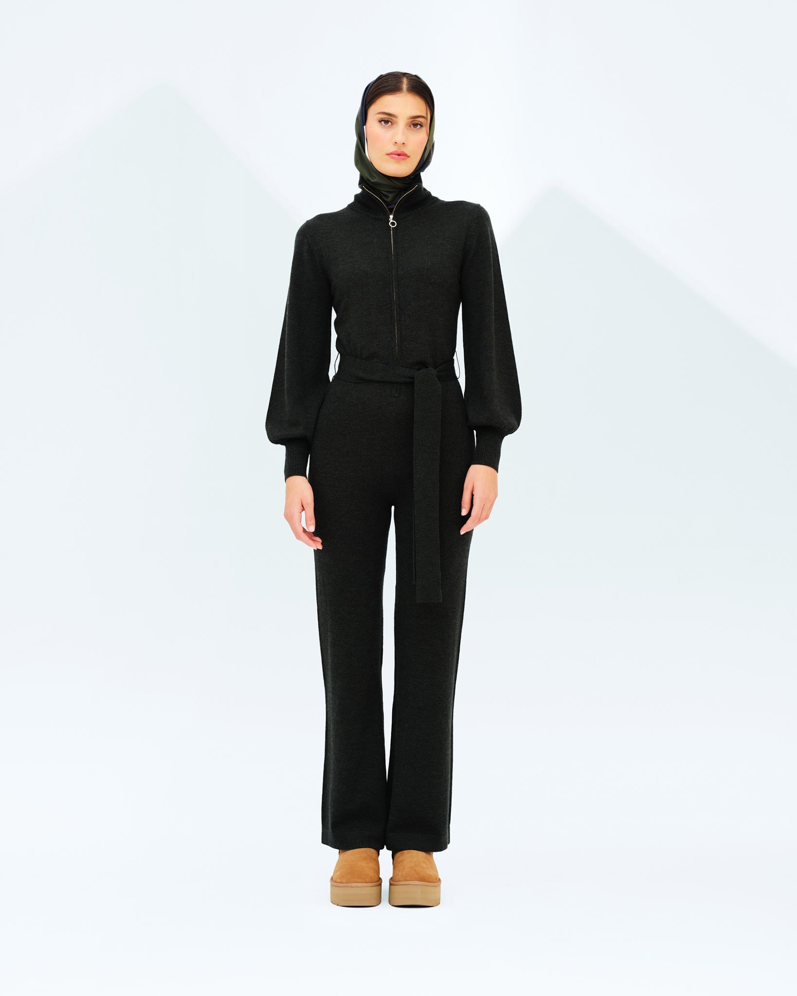 A woman models the sleek Geilo Jumpsuit Women by We Norwegians, featuring a black long-sleeved design with a high collar and a matching belt. Standing against a white background, she accessorizes with a headscarf and light brown platform shoes. The minimalist setting highlights her outfit, evoking the elegance of a chic mountain resort.