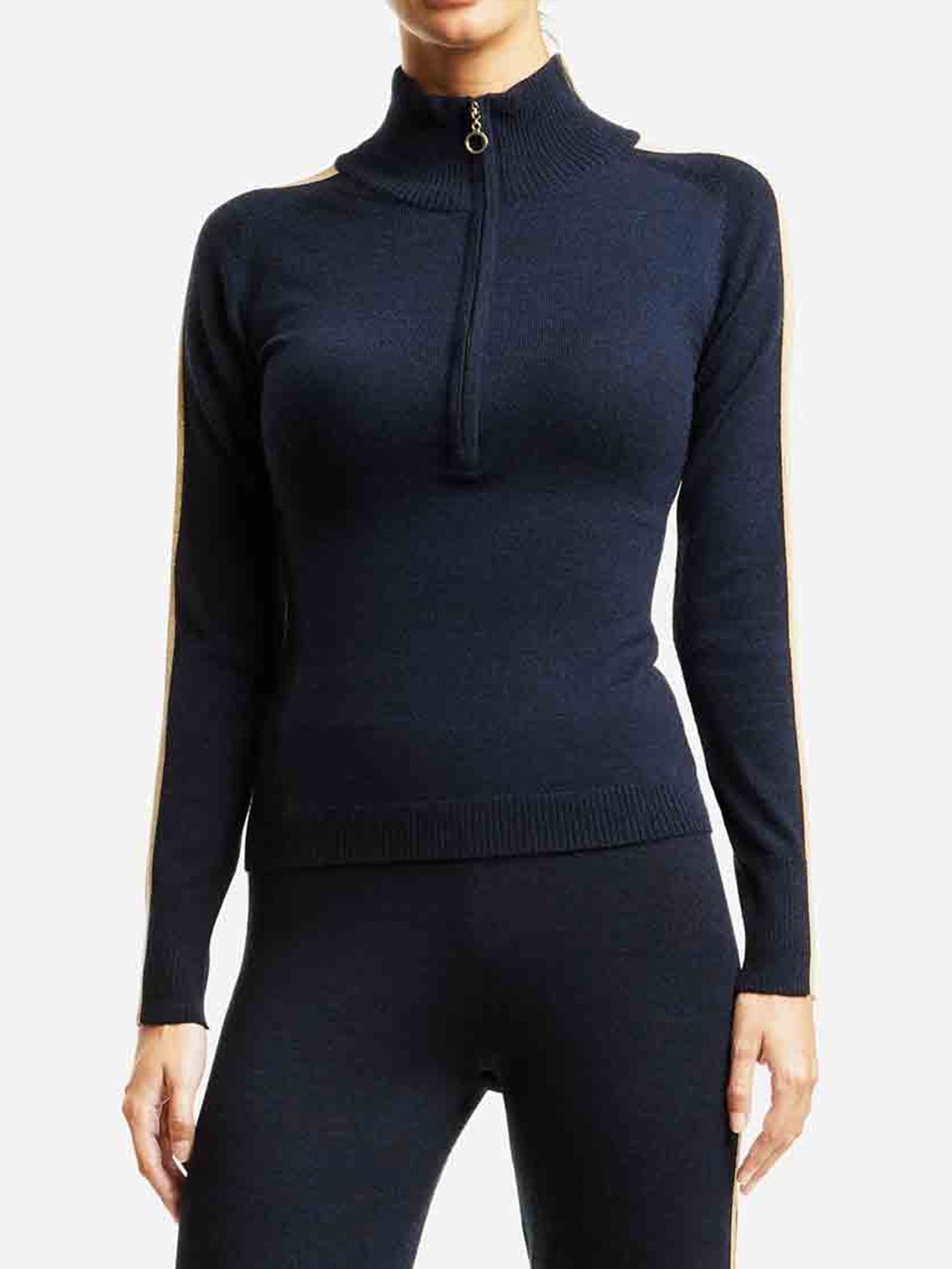 A person wearing a sleek, long-sleeved, black Geilo Zip Up Women top by We Norwegians, with a high collar and matching black leggings. The top, possibly crafted from Merino wool, features a gold accent stripe running down the sleeves. The outfit is simple yet elegant, highlighting a form-fitting and comfortable style perfect for après ski.