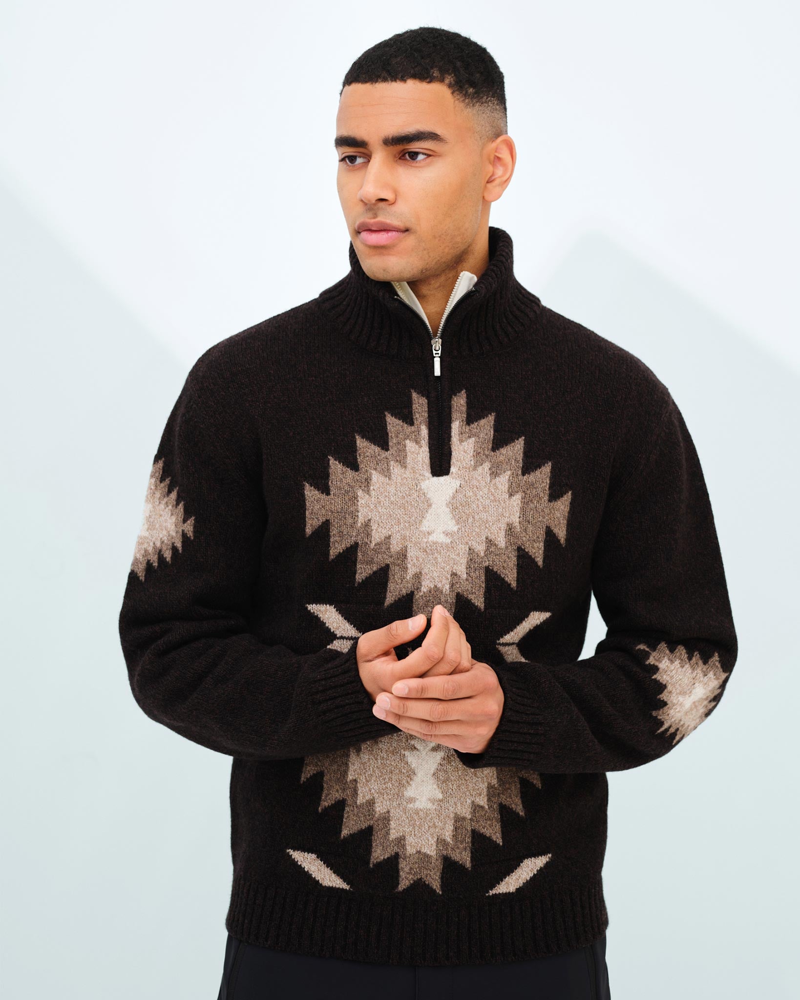 A man stands against a light background, wearing the Hovden Sweater Men by We Norwegians. This black knitted wool sweater features beige geometric patterns and a convenient quarter-zip collar. With short hair and a pensive expression, he clasps his hands together in front of him, perfectly capturing the essence of Fall weather.