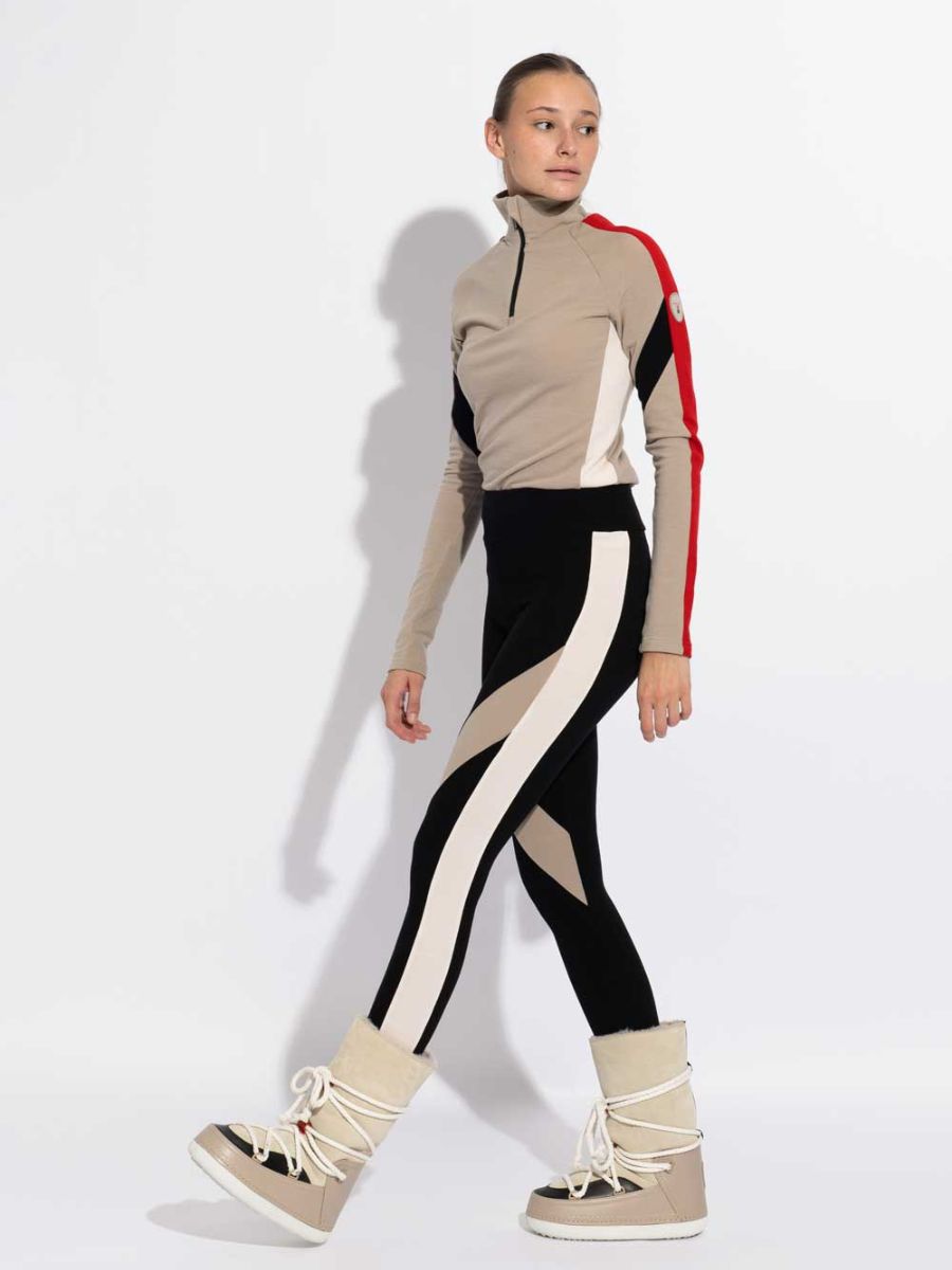 A person stands against a white background wearing an exclusive capsule collection sports outfit from We Norwegians. The outfit features a beige and black long-sleeve top with red accents paired with matching leggings adorned with geometric patterns. They complete the look with INUIKII + WN Zip Up Women lace-up boots in beige, featuring thick soles and high, padded tops.