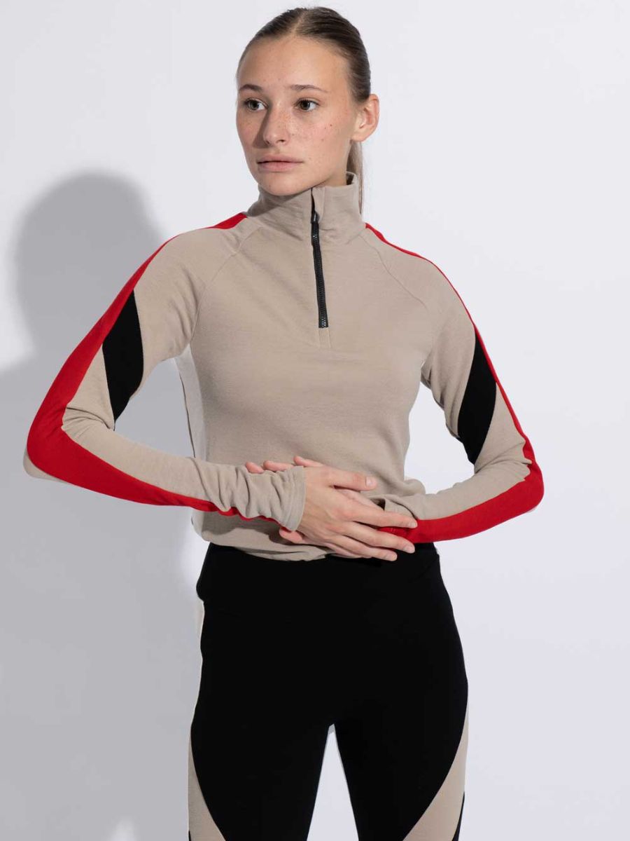 An individual with long hair tied back sports the INUIKII + WN Zip Up Women from designer brand We Norwegians. This light beige zip-up top features red and black accents on the sleeves, forming part of a designer capsule collection. Complementing the look are black and beige pants. The person stands with hands clasped at their waist against a neutral backdrop, gazing off to the side.