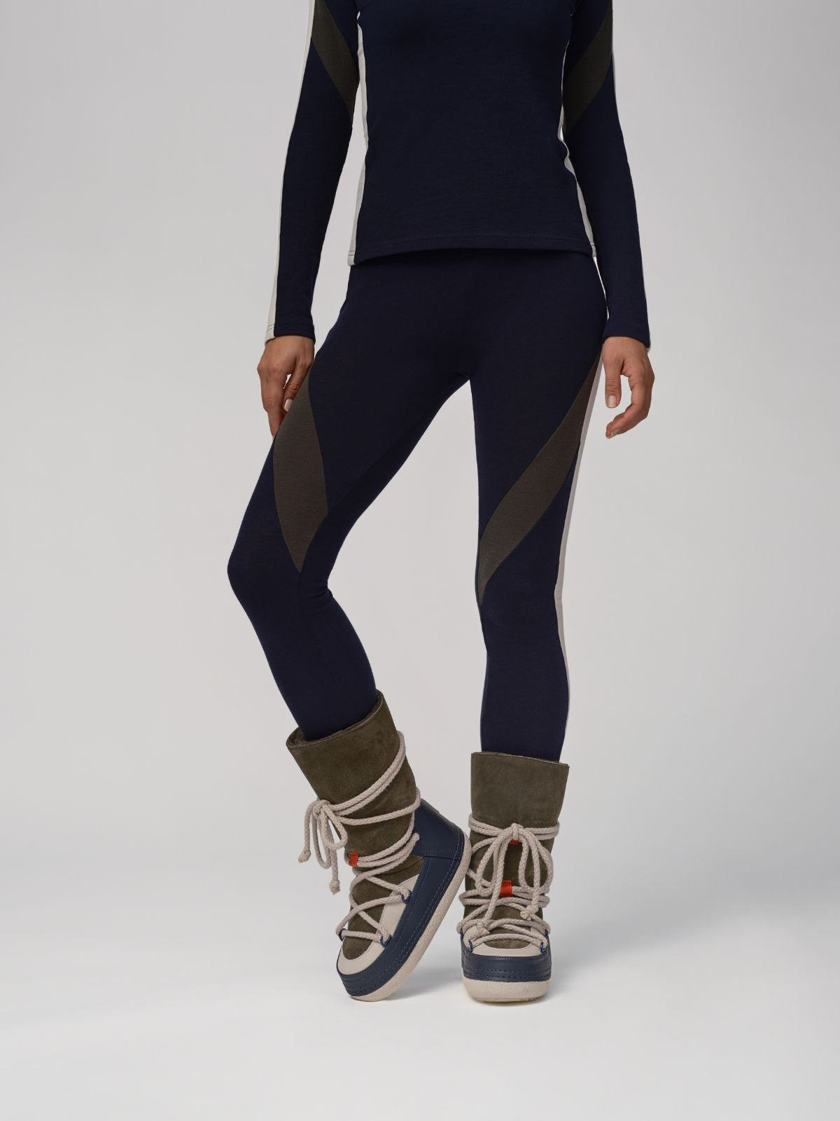 The person is dressed in a navy and olive activewear set, featuring We Norwegians Inuikii+WN Leggings Women made from Merino wool with long sleeves. Paired with high-top winter boots that boast laces and a thick sole, they're perfectly styled for ski fashionistas against a plain background.