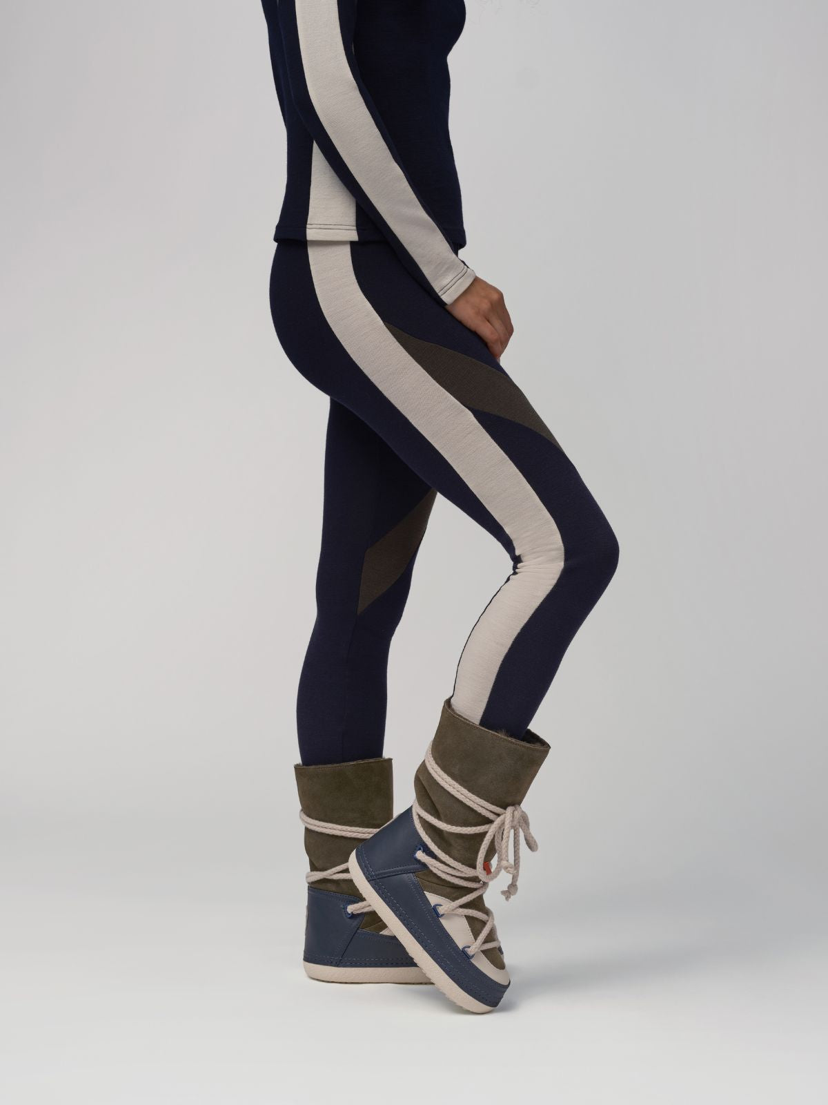 A person showcases their ski fashionista flair by wearing the We Norwegians Inuikii+WN Leggings Women, paired seamlessly with a navy and cream striped top and blue and green lace-up boots. The image highlights their stylish lower half against a plain backdrop.