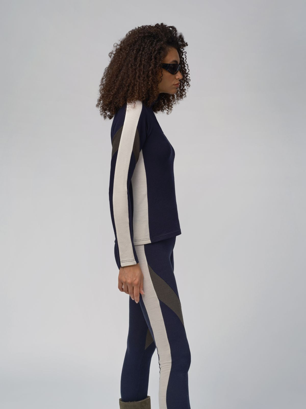 A woman with curly hair stands in profile against a plain backdrop. She is wearing the INUIKII + WN Zip Up Women by We Norwegians, a form-fitting, long-sleeve top and matching leggings featuring contrasting panels of dark blue and beige from a designer capsule collection. She also wears dark sunglasses and green boots.