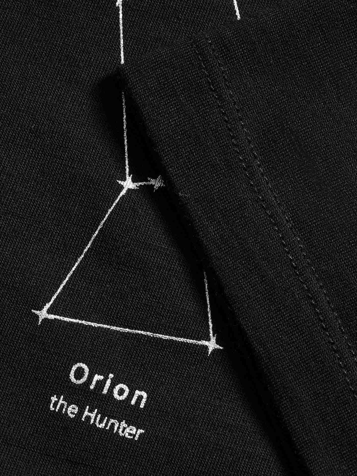 Close-up of a black fabric from the limited edition Natt LongSleeve PJ Women Black by We Norwegians, featuring a white, minimalist outline of the Orion constellation. Below it is stitched text reading "Orion the Hunter." Some details of this luxurious pajama design are partially obscured by an overlapping piece of the same fabric.