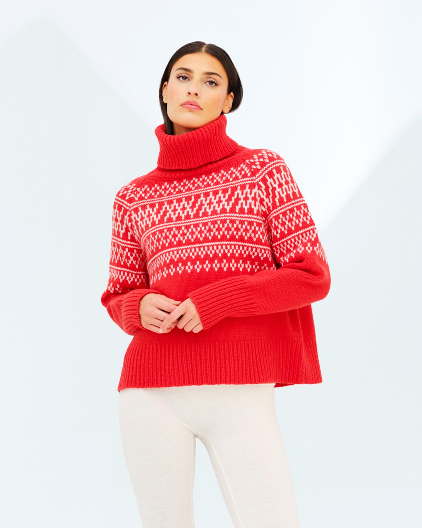 A person with long, dark hair is wearing a red We Norwegians Setesdal Sweater Women, featuring white geometric patterns and crafted from luxurious merino cashmere yarn. They are also dressed in light-colored pants and are standing against a light background while looking slightly upward.