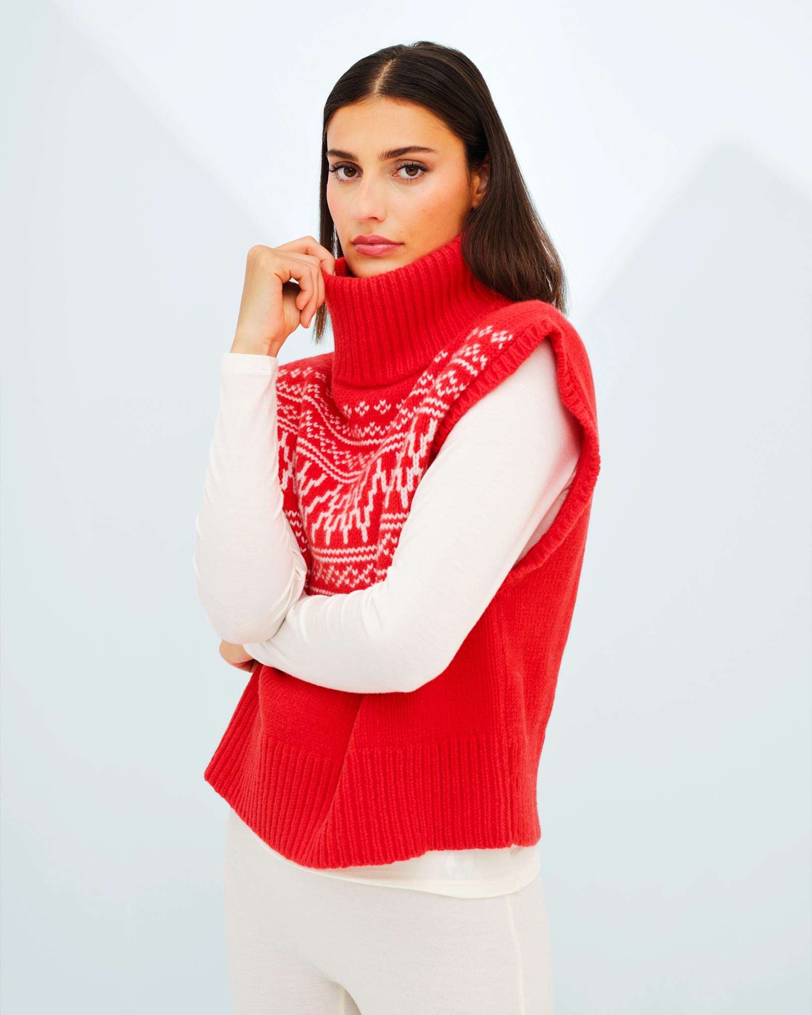 A person with long dark hair is wearing the We Norwegians Setesdal Vest Women, a red sleeveless turtleneck sweater featuring an intricate white pattern on the chest reminiscent of Norwegian knits. They have a white long-sleeved top underneath and light-colored pants, standing against a light, blurred background.