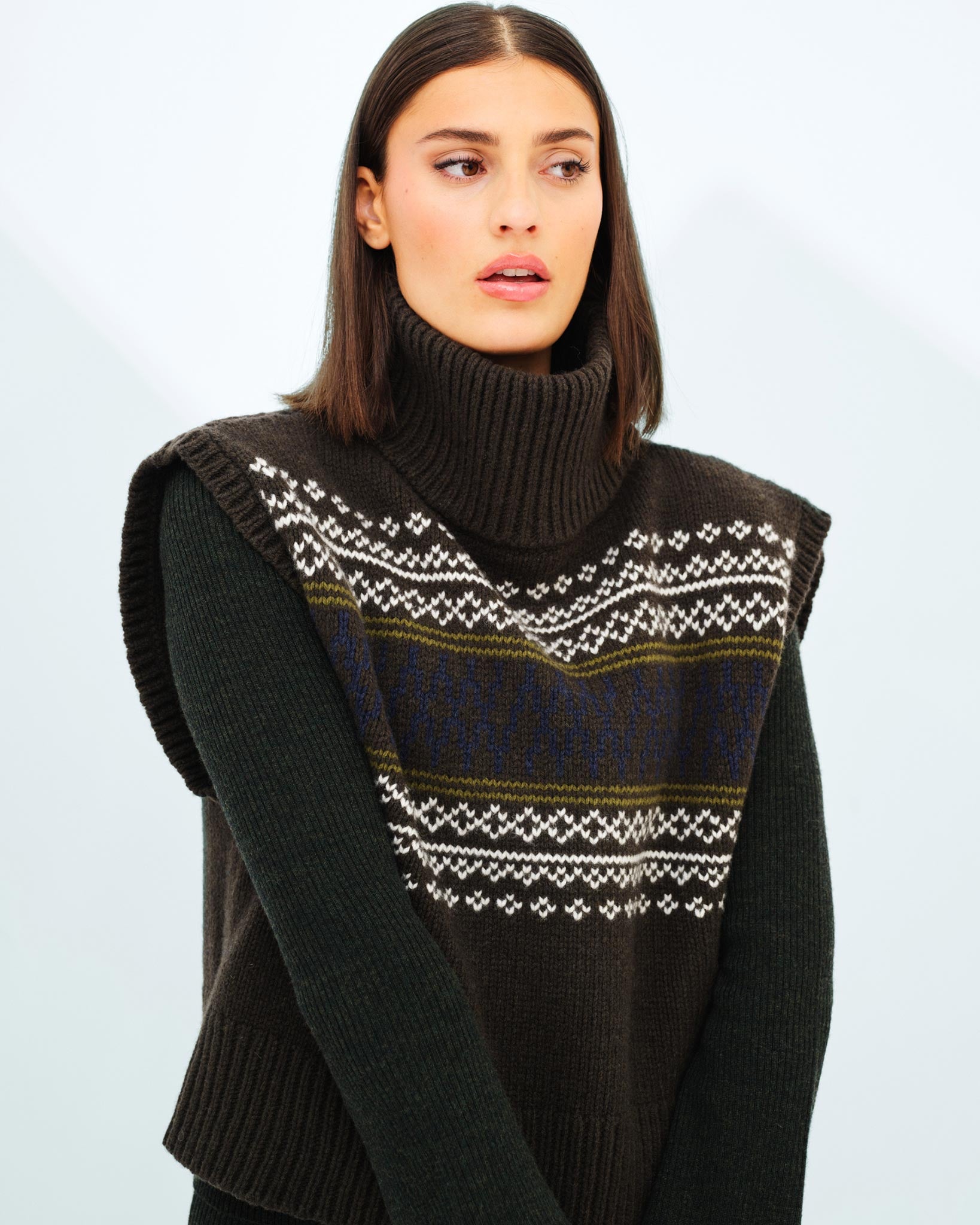 A woman with shoulder-length dark hair wears a dark turtleneck sweater paired with the Setesdal Vest Women by We Norwegians. She is looking slightly to the side with a neutral expression while standing against a plain, light-colored background.