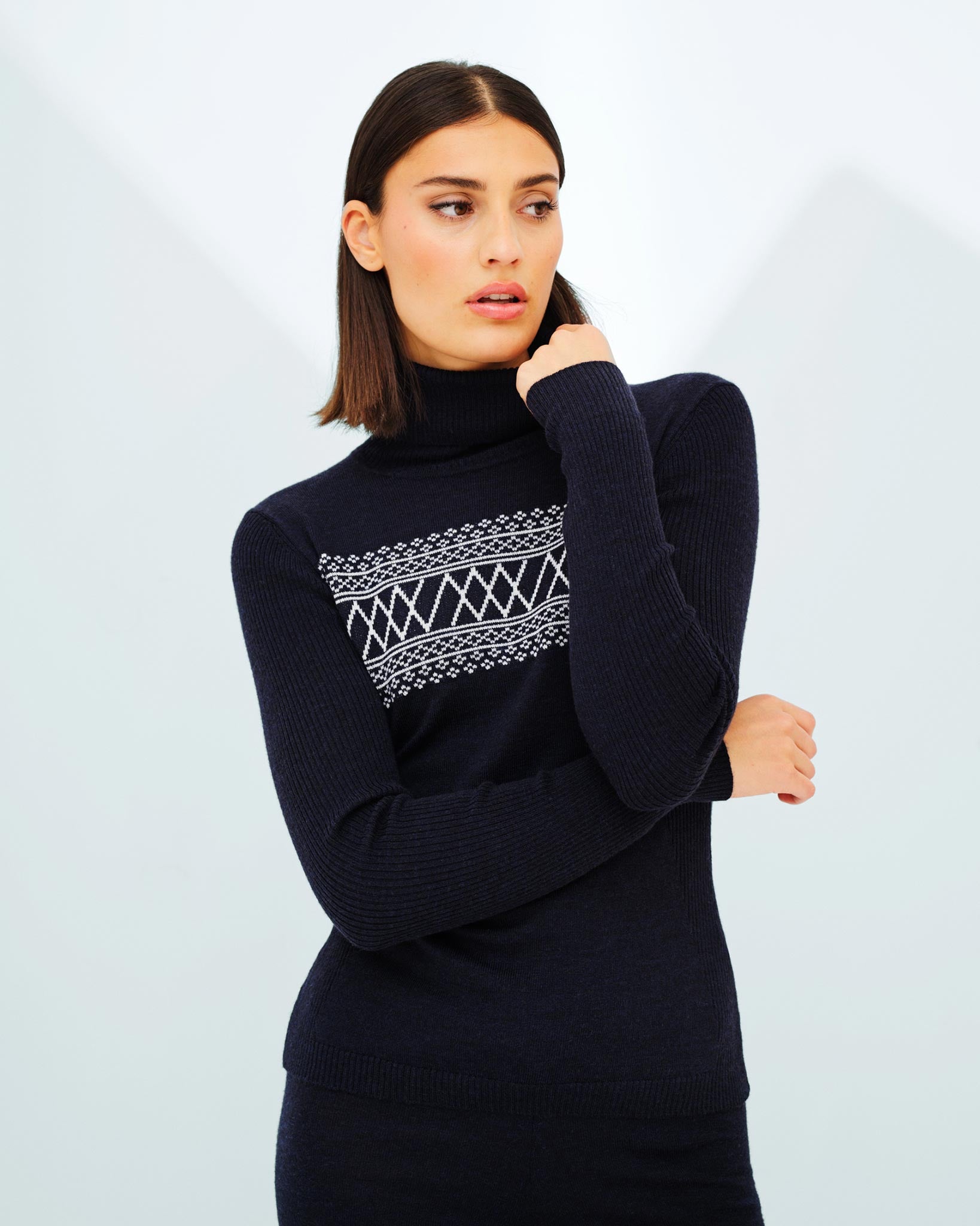 A person with shoulder-length brown hair wears the Signature Rollneck Women by We Norwegians, featuring a white geometric pattern across the chest. They have one hand on their forearm and are looking to the side against a plain background, giving off retro ski outfit vibes.